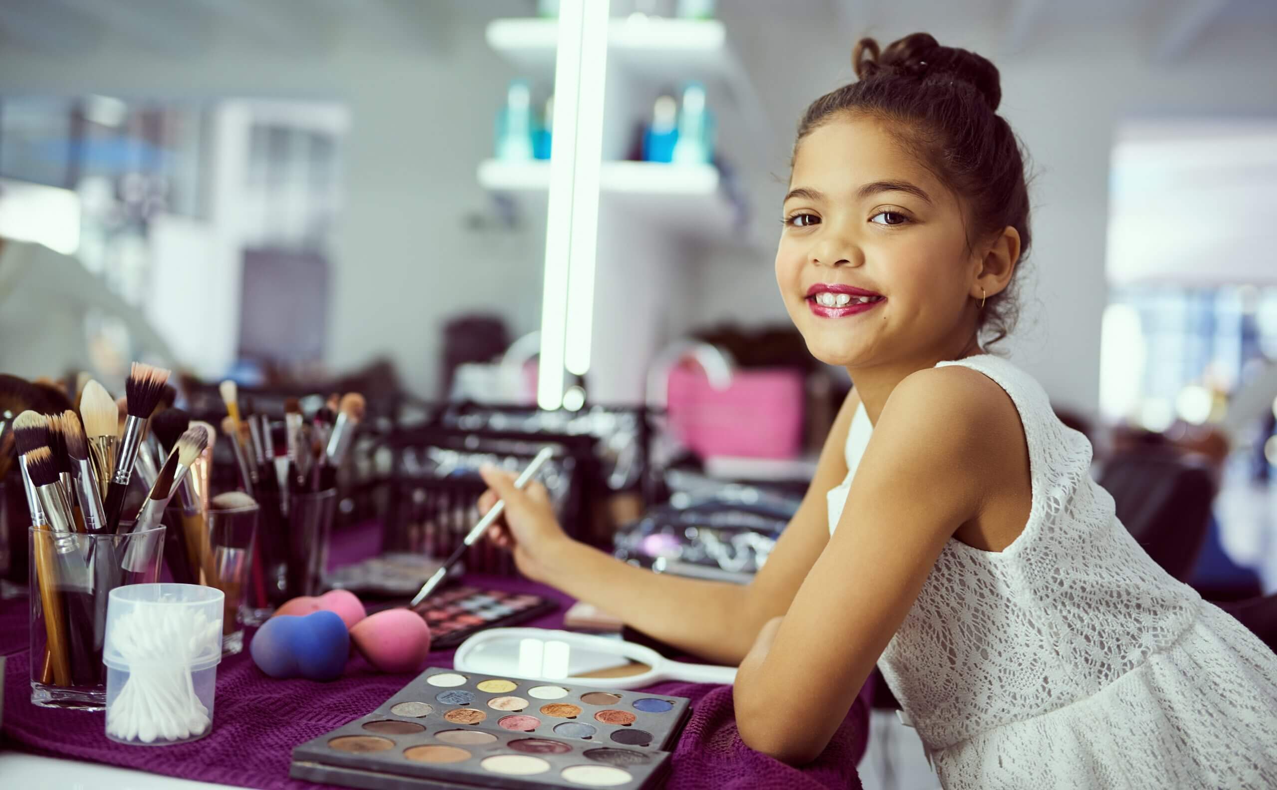 Pageant Child standing at a makeup table smiling at the camera. She is wearing makeup with her hair up and a pretty white dress on.